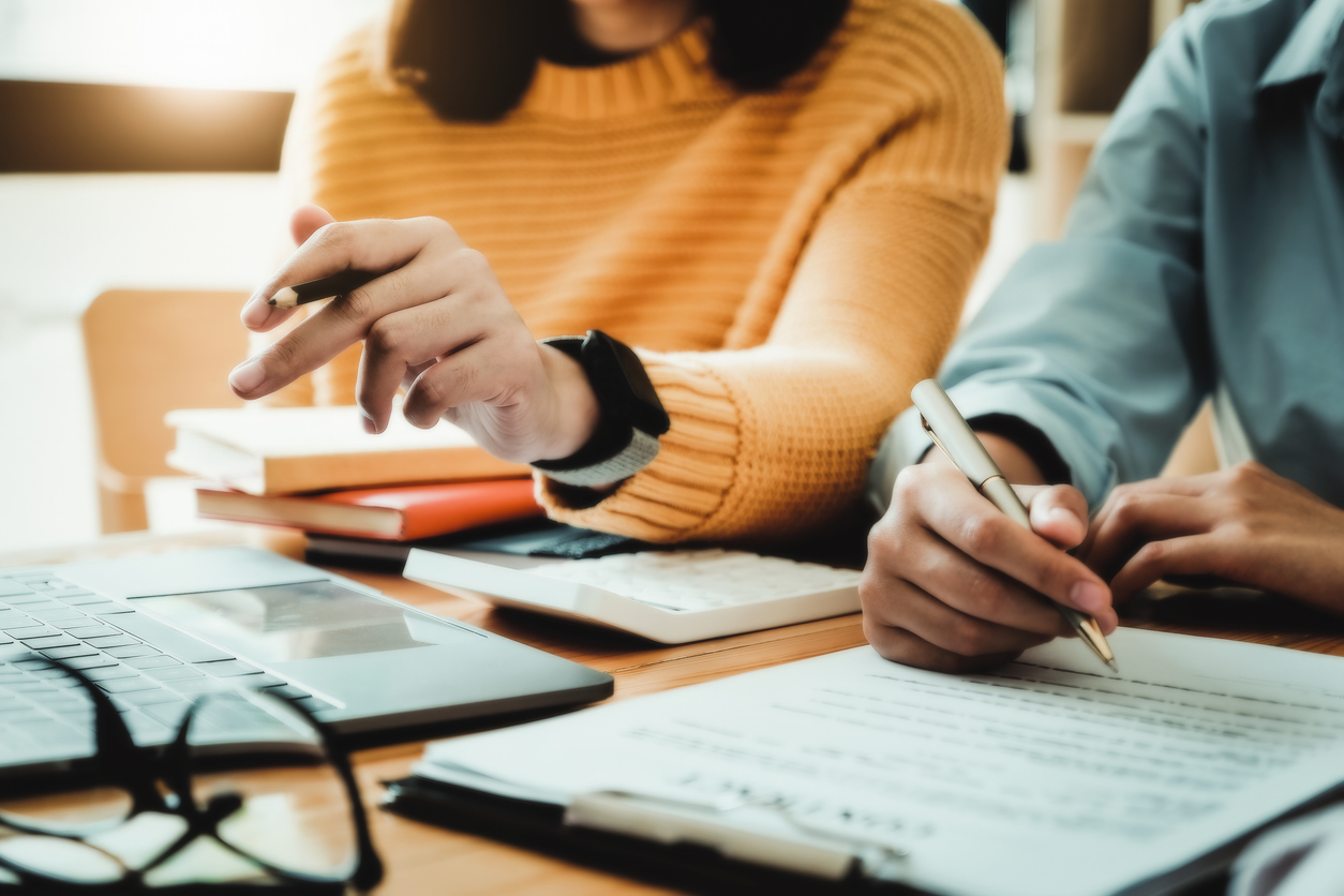 Emphasis is on the yellow-shirted consultant holding a pencil and the business owner holding a pen reading the terms of a joint venture contract with a partner company.