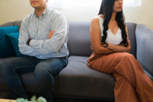 Upset couple sitting with arms crossed on a couch during couples therapy session with psychologist, having relationship problems and feeling angry